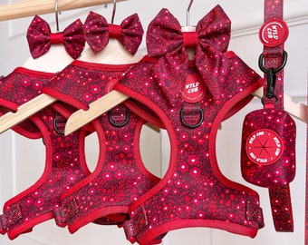 May include: Three red dog harnesses with floral patterns and matching red bow ties. The harnesses have adjustable straps and a clip closure. The bow ties are attached to the harnesses with a velcro strap. The harnesses are hanging on wooden hangers. The text "NYLD CUB" is printed on the harnesses.