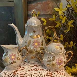 May include: A white porcelain tea set with gold accents and floral designs. The set includes a teapot, creamer, and sugar bowl.