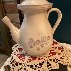 May include: A cream-colored ceramic teapot with a blue and red floral design. The teapot has a lid and a handle. It is sitting on a book with a crocheted doily.