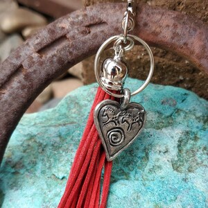 May include: A red tassel keychain with a silver heart charm featuring a design of three horses. The charm is attached to a silver ring with a silver bell.