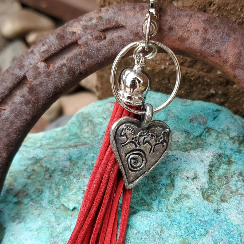 May include: A red tassel keychain with a silver heart charm featuring a design of three horses. The charm is attached to a silver ring with a silver bell.
