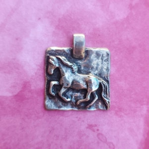 May include: Silver pendant featuring a detailed, textured horse design. The pendant is rectangular and has a rustic, aged look.