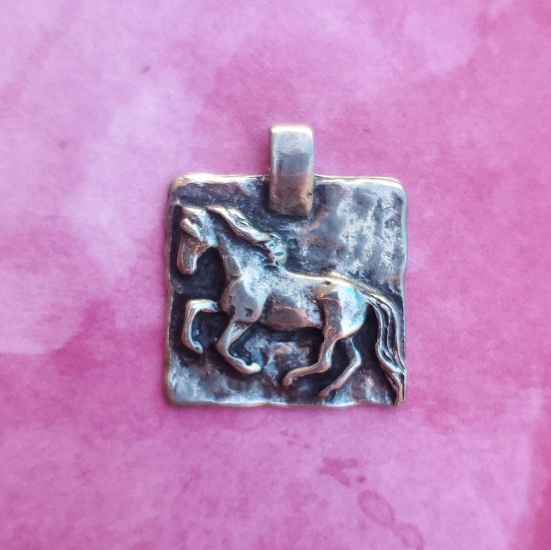 May include: Silver pendant featuring a detailed, textured horse design. The pendant is rectangular and has a rustic, aged look.