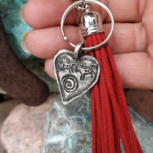 May include: Silver heart-shaped keychain with a design of three horses and a spiral. The keychain has a red tassel.