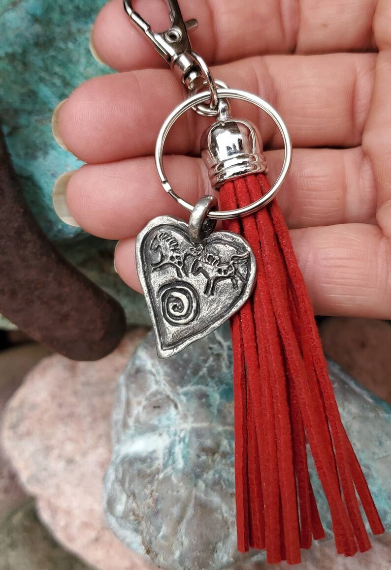 May include: Silver heart-shaped keychain with a design of three horses and a spiral. The keychain has a red tassel.