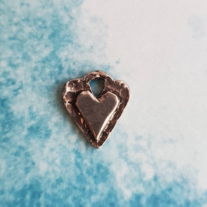 May include: A small, silver-colored heart-shaped pendant with a textured border and a smaller heart in the center. The pendant has a loop at the top for attaching to a chain or cord. The background is a light blue and white pattern.