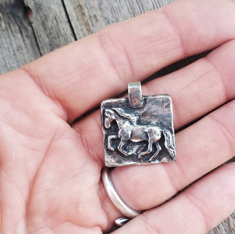 May include: A silver pendant featuring a detailed, three-dimensional sculpture of a horse running. The pendant is square-shaped and has a textured surface.