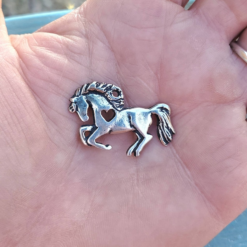 May include: Silver charm of a horse with a heart cut out of its side. The horse is running with its mane flowing.