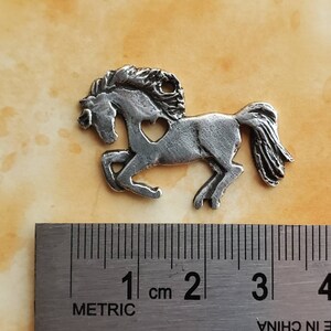 May include: A silver-colored metal charm of a horse with a heart cut out of its side. The charm is approximately 2 cm long.