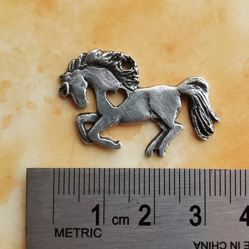 May include: A silver-colored metal charm of a horse with a heart cut out of its side. The charm is approximately 2 cm long.