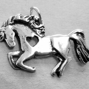 May include: Silver charm of a horse with a heart cut out of its body. The horse is running with its mane flowing in the wind.