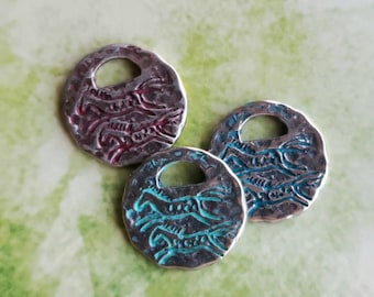 May include: Three silver-toned, round pendants with a hammered texture and a small hole at the top. Each pendant features a stylized horse design in red or teal. The pendants are displayed on a green and white surface.