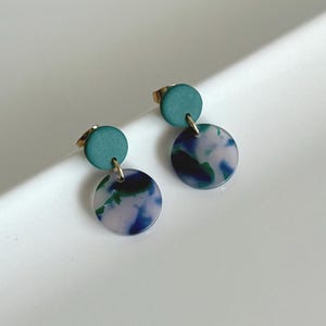 Könnte beinhalten: Ein Paar Ohrringe mit einem teal-farbenen oberen Kreis und einem größeren, marmorierten Kreis in Blau, Grün und Weiß. Die Ohrringe haben goldfarbene Stifte und sind vor einem weißen Hintergrund platziert.