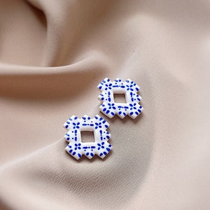 May include: A pair of square earrings featuring a white base with a blue floral pattern. The earrings are set against a beige fabric backdrop. Each earring has a geometric design with a square centre.