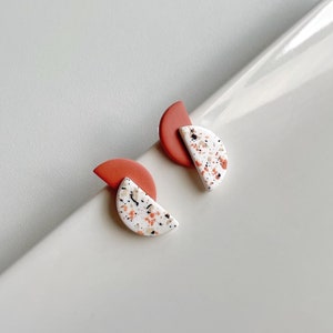 Pode incluir: Um par de brincos geométricos com um design branco e vermelho salpicado. Os brincos são feitos de argila polimérica e têm a forma de um semicírculo.