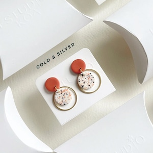 Puede incluir: Un par de pendientes de aro dorados con un pequeño disco de arcilla moteado de naranja y blanco unido a cada aro. Los pendientes se muestran en una tarjeta blanca con el texto "GOLD & SILVER" impreso en ella. La tarjeta está encima de un embalaje de almohada blanco con el texto "STUDIO KOA" en relieve.