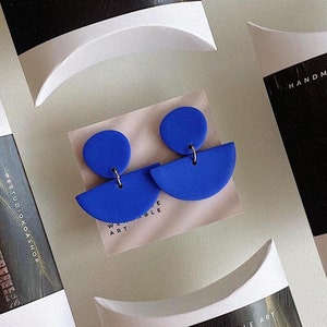 K&ouml;nnte beinhalten: Zwei Paar blaue Polymer Clay Ohrringe in Halbkreisform. Die Ohrringe sind in wei&szlig;en Kissenboxen mit schwarzen und wei&szlig;en Etiketten verpackt, auf denen "Handmade Wearable Art" und "Studio Koa Shop" steht.