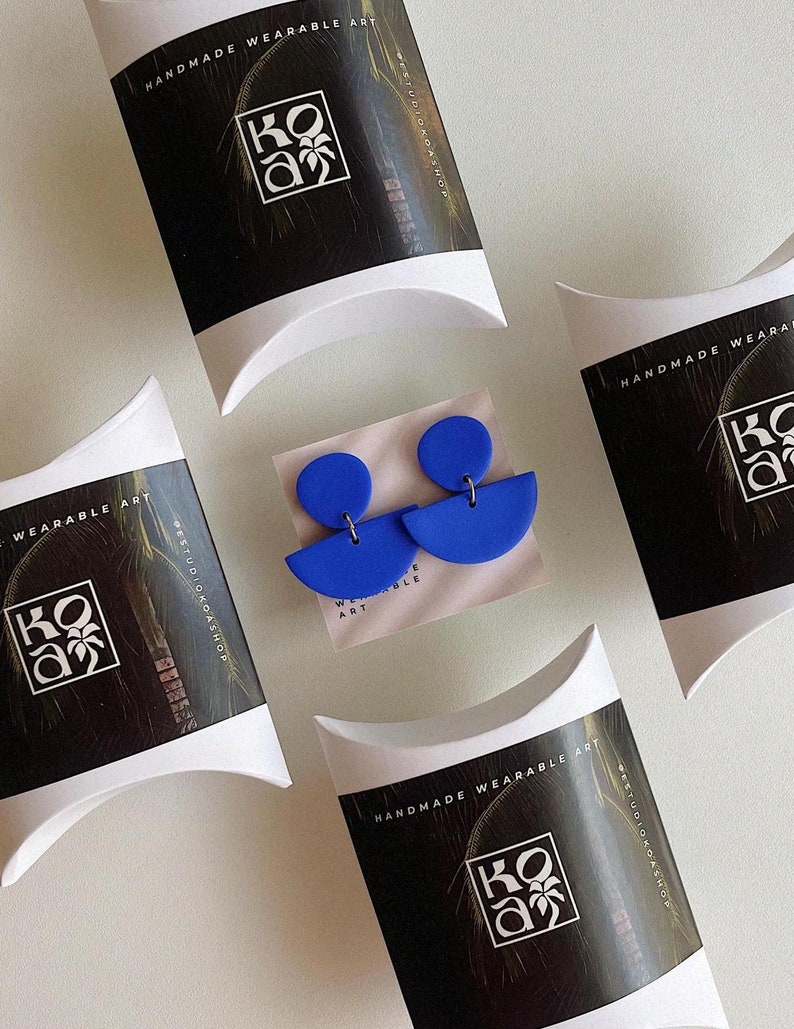 K&ouml;nnte beinhalten: Zwei Paar blaue Polymer Clay Ohrringe in Halbkreisform. Die Ohrringe sind in wei&szlig;en Kissenboxen mit schwarzen und wei&szlig;en Etiketten verpackt, auf denen "Handmade Wearable Art" und "Studio Koa Shop" steht.