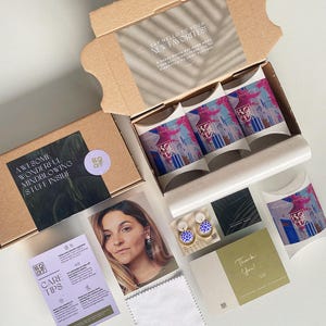 May include: A cardboard box, open to reveal packaged products, including three boxes with colourful designs. Other items include a photo, care tips, a thank you card, and a cleaning cloth. Text includes "Say Hello to Your New Favorites!" and "Awesome Wonderful Mindblowing Stuff Inside."