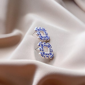 May include: A pair of white stud earrings with a square, openwork design. Each earring features a repeating pattern of small blue floral details. The earrings are set against a soft, neutral-toned fabric background.