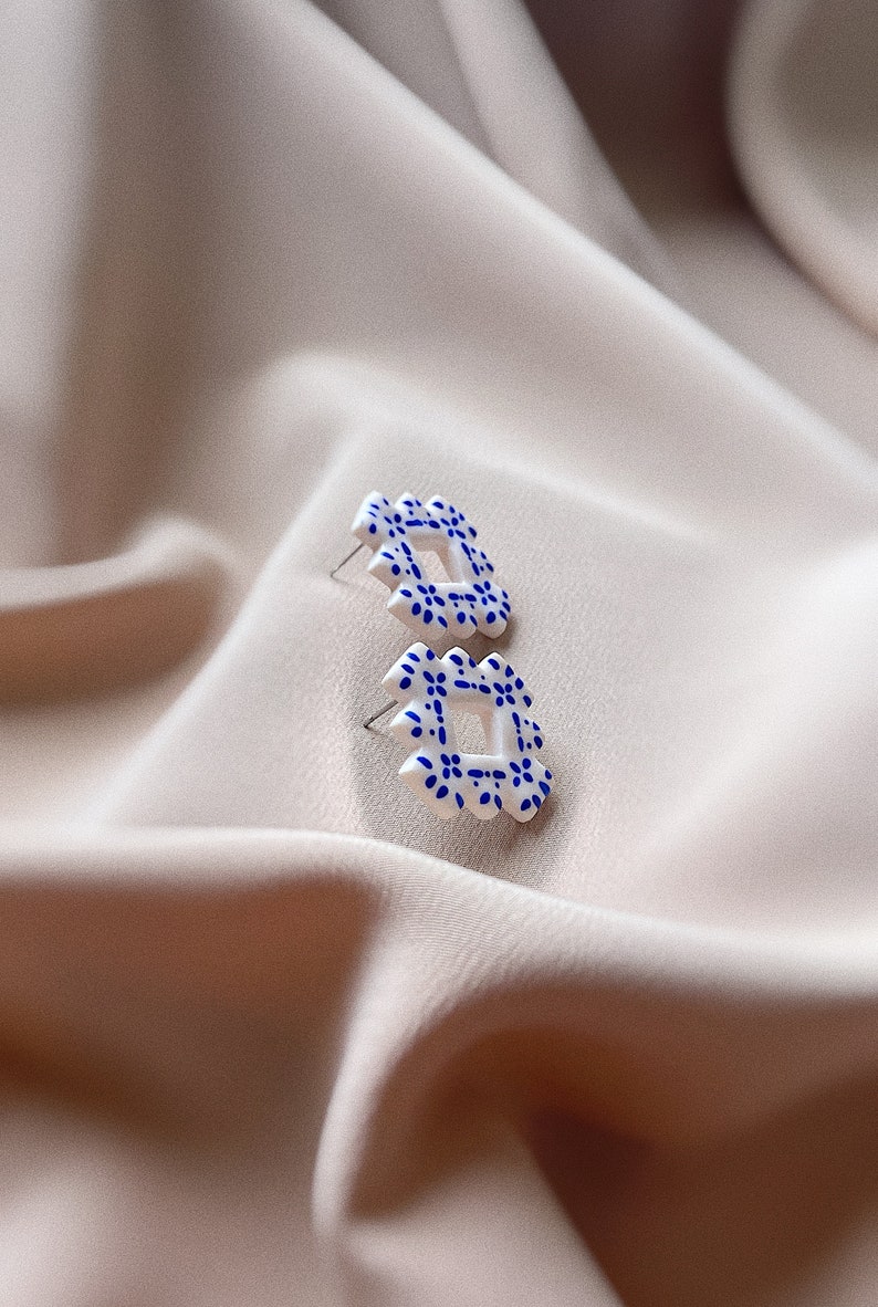 May include: A pair of white stud earrings with a square, openwork design. Each earring features a repeating pattern of small blue floral details. The earrings are set against a soft, neutral-toned fabric background.