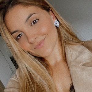 May include: A close-up of a person wearing white and silver stud earrings with a floral design. The person has long blonde hair and is wearing a tan jacket. The earrings are the focal point.