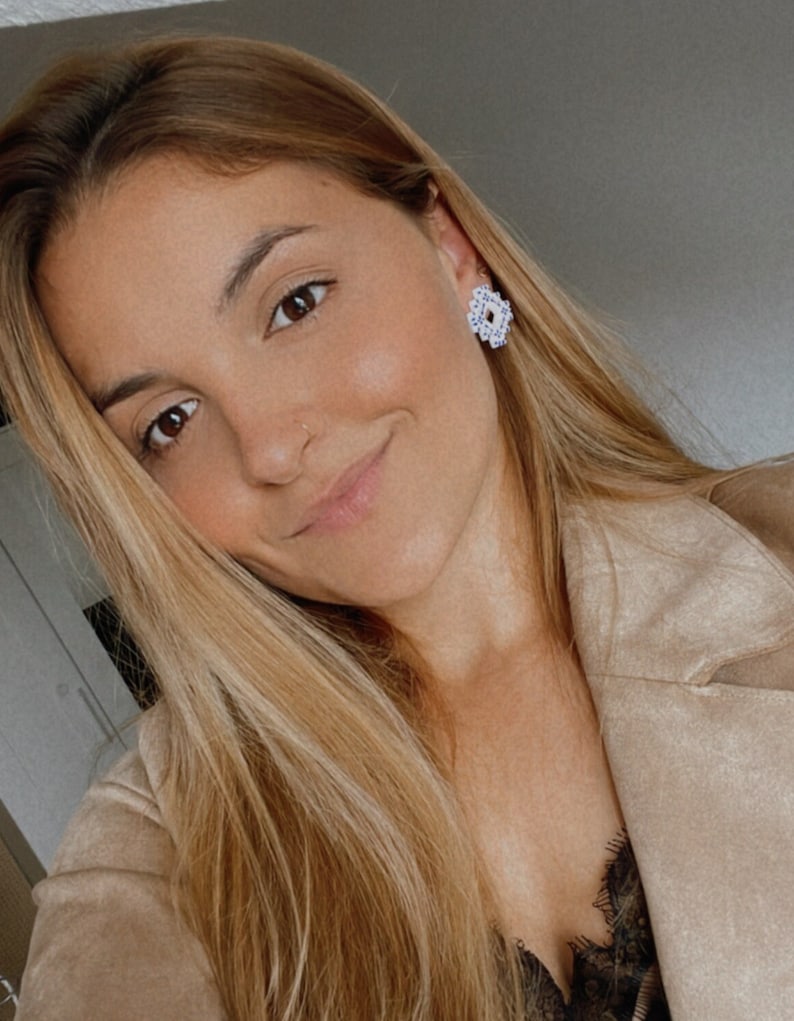 May include: A close-up of a person wearing white and silver stud earrings with a floral design. The person has long blonde hair and is wearing a tan jacket. The earrings are the focal point.
