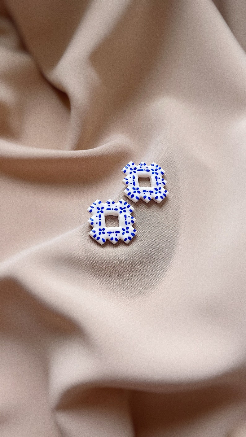 May include: A pair of square earrings featuring a white base with a blue floral pattern. The earrings are set against a beige fabric backdrop. Each earring has a geometric design with a square centre.