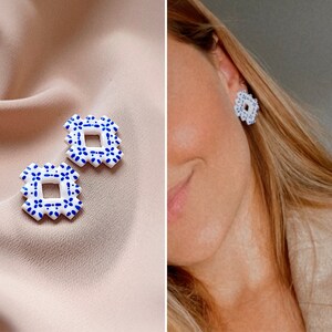 May include: White and blue square earrings with a floral pattern. The earrings are displayed on a beige surface and also worn. The earrings feature a geometric design with a square centre and a scalloped edge.