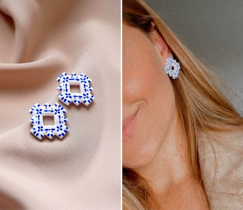 May include: White and blue square earrings with a floral pattern. The earrings are displayed on a beige surface and also worn. The earrings feature a geometric design with a square centre and a scalloped edge.