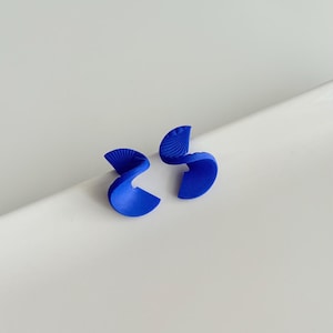 Op de afbeelding: Twee kobaltblauwe oorbellen met een abstract, lintachtig ontwerp. De oorbellen hebben getextureerde details en worden gepresenteerd op een witte ondergrond. Het ontwerp is modern en opvallend.