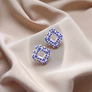 May include: Two white square earrings with a blue floral pattern. The earrings are displayed on a soft, beige fabric. The earrings have a geometric shape with a repeating pattern of small blue flowers.