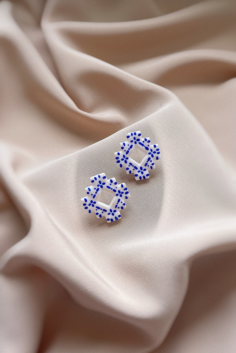 May include: Two white square earrings with a blue floral pattern. The earrings are displayed on a soft, beige fabric. The earrings have a geometric shape with a repeating pattern of small blue flowers.
