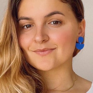K&ouml;nnte beinhalten: Eine Frau mit langen blonden Haaren tr&auml;gt ein wei&szlig;es Hemd und eine goldene Kettenkette. Sie tr&auml;gt blaue geometrische Ohrringe.