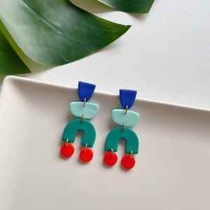 Op de afbeelding: Geometrische oorhangers in blauw, teal en rood. De oorbellen hebben een gelaagd ontwerp met een blauw bovenstuk, een teal middengedeelte en een teal boog met een rode cirkel aan de onderkant. De oorbellen worden gepresenteerd op een witte ondergrond.