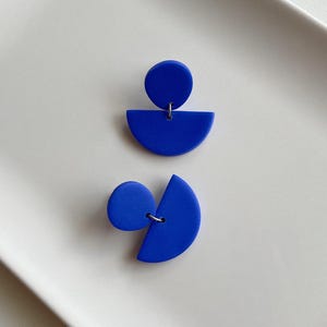 K&ouml;nnte beinhalten: Ein Paar blaue geometrische Ohrringe. Jeder Ohrring besteht aus einem Kreis und einer Halbkreisform.