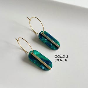 May include: A pair of gold hoop earrings with teal and green marbled oval pendants. Each pendant has a gold-colored bar across the center. The text "GOLD & SILVER" is visible in the image.