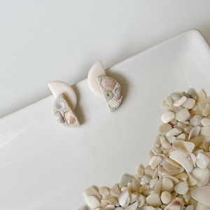 Peut inclure: Une paire de boucles d'oreilles de couleur crème en forme de demi-cercle. Les boucles d'oreilles sont décorées de petites coquillages dans différentes nuances de beige, rose et brun.