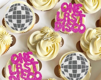 May include: A box of cupcakes decorated with a disco ball design and the words "One Last Disco" in pink glitter. The cupcakes are frosted with white frosting and have silver sprinkles.