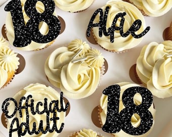 May include: A box of cupcakes decorated with black glitter frosting and various toppers. The toppers include the numbers "18", the words "Official Adult", and the name "Alfie".