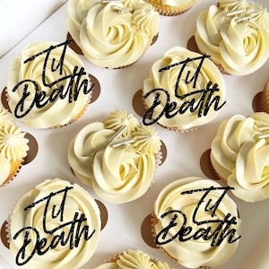 May include: A box of cupcakes with white buttercream frosting, each decorated with a black glitter "Til Death" cake topper. The cupcakes are in brown paper cases and have silver sprinkles.