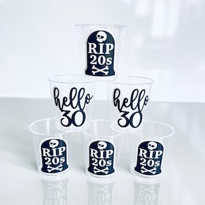 May include: Clear plastic shot glasses with black text and graphics. Some glasses say "RIP 20s" with a tombstone and crossbones, while others say "hello 30". The shot glasses are stacked on top of each other.