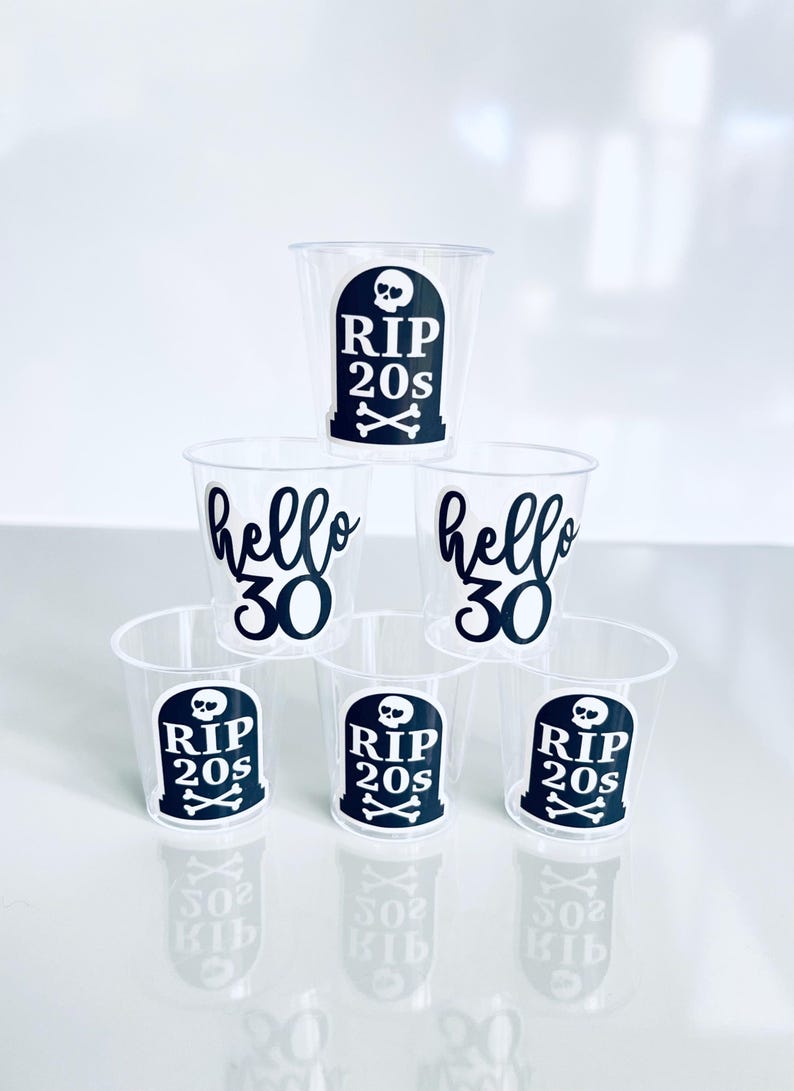 May include: Clear plastic shot glasses with black text and graphics. Some glasses say "RIP 20s" with a tombstone and crossbones, while others say "hello 30". The shot glasses are stacked on top of each other.