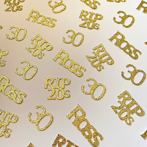 May include: Gold glitter confetti with the phrases "RIP 20s", "30", and "Boss" scattered across a white surface. The confetti is in a variety of sizes and orientations, creating a celebratory theme.
