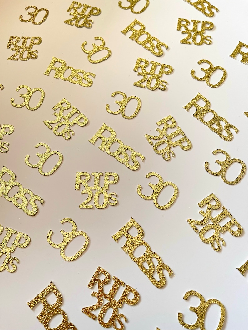 May include: Gold glitter confetti with the phrases "RIP 20s", "30", and "Boss" scattered across a white surface. The confetti is in a variety of sizes and orientations, creating a celebratory theme.