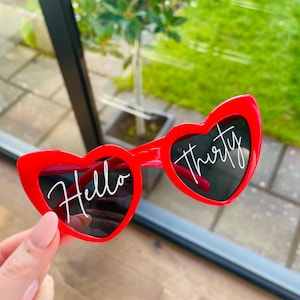 May include: Red heart-shaped sunglasses featuring the white script text Hello Thirty printed across the dark lenses. The frames are bright red with a smooth finish, and the lenses are dark, providing a clear contrast for the white lettering.