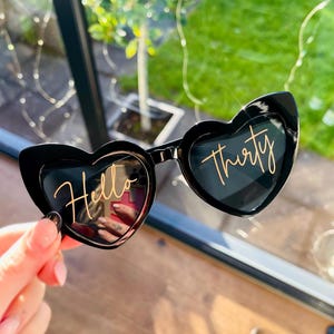 May include: Black heart-shaped sunglasses featuring the gold cursive text Hello Thirty printed across the lenses. The frames are made of a glossy black material and have a distinct heart silhouette, with the word Hello on the left lens and Thirty on the right lens.