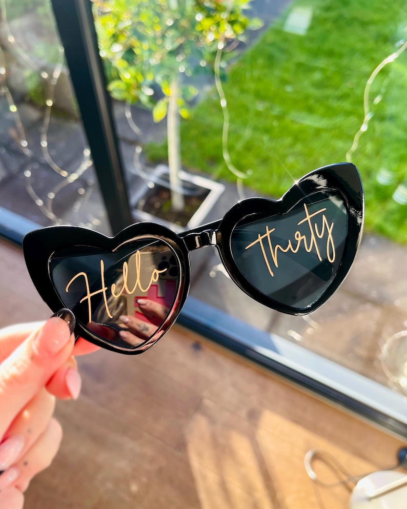 May include: Black heart-shaped sunglasses featuring the gold cursive text Hello Thirty printed across the lenses. The frames are made of a glossy black material and have a distinct heart silhouette, with the word Hello on the left lens and Thirty on the right lens.