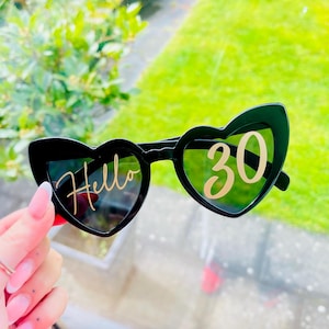 May include: Black heart-shaped sunglasses featuring gold lettering on the lenses. The left lens displays the word Hello in a cursive script, while the right lens features the number 30 in a bold, sans-serif font. The frames are solid black plastic.