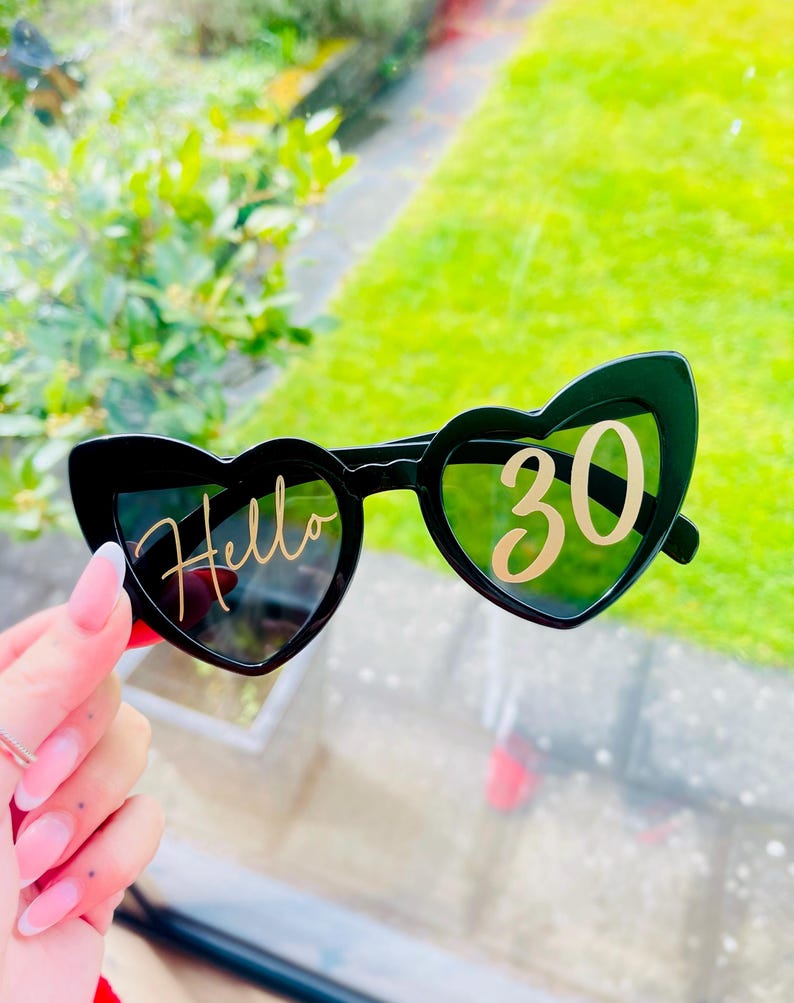 May include: Black heart-shaped sunglasses featuring gold lettering on the lenses. The left lens displays the word Hello in a cursive script, while the right lens features the number 30 in a bold, sans-serif font. The frames are solid black plastic.